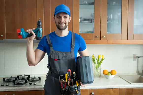 Repairman holds a screwdriver and a suitcase of tools in the kitchen and looks at the camera